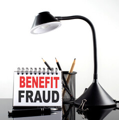 BENEFIT FRAUD text on notebook with pen and table lamp on the black background
