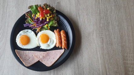 Top view of fried egg and sausage and ham smiley face and colorful salad like hair on black plate at wood table, breakfast serve for kid, funny breakfast.