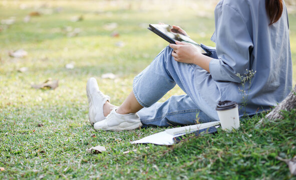 Young asian woman with digital tablet sits on the grass in the park. she using tablet online working, remote work and communication in social networks and working part time and relaxing 