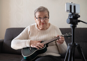 Elderly woman 70+ sits on the couch and learns to play the ukulele at home using video tutorial on her smartphone. Adaptation of pensioners in the modern world. New knowledge and skills. Hobby