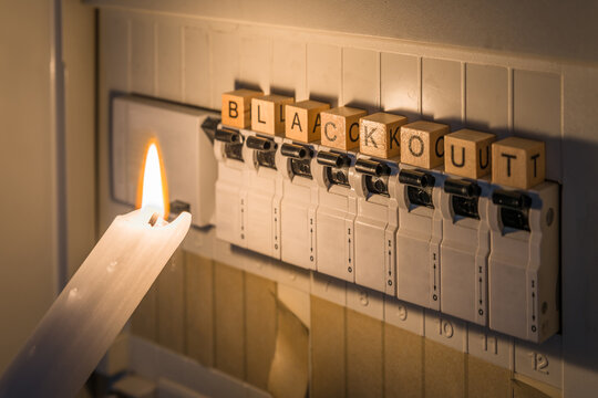 Sicherungskasten mit Sicherungen in einem Verteilerkasten während eines Stromausfall mit weißer Kerze beleuchtet die ein Mann in der Hand hält mit dem Wort Blackout als Text, Deutschland