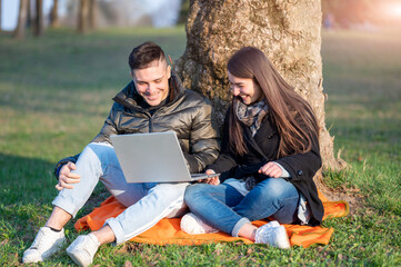 A beautiful couple in casual wear is having fun with the laptop outdoors. Sitting at the foot of a tree, you enjoy nature on a sunny spring day. Remote teamwork concept.