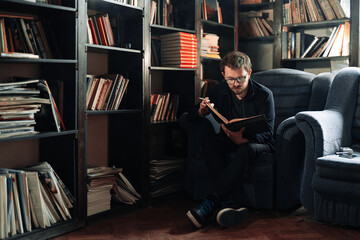 Adult student reading a book in the college library while smoking. Young male wearing glasses with bookshelves on background sitting on couch.