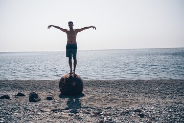 Man doing yoga exercise outdoors standing on old rusty floating marine mine on the beach with rocky...