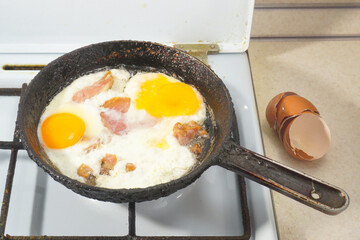 Fried eggs with pieces of meat are fried on a gas stove in a black frying pan.