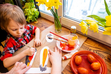 women's speckled hands teach boy draws paper rabbit ears. Preparation for Easter: a child paints paper rabbit ears with paints in the kitchen among fresh greenery, flowers and painted eggs