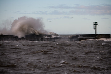 Olbrzymi sztormowy bałwan morski rozbija się o falochron. Widać ślady zniszczenia części falochronu przez żywioł morski.