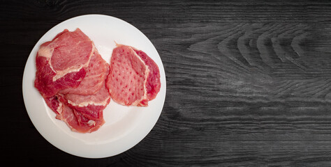 Raw pork chop on a white plate on a black wooden background. Fatty meat beaten off for cooking.