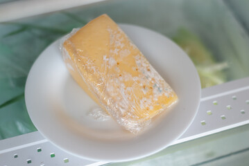 Moldy cheese wrapped in cling film in the refrigerator. Incorrect storage, spoiled product, expired concept.