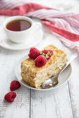 A slice of puff pastry pie with raspberries on a plate, a cup of coffee and a towel. Delicate and airy composition, Napoleon pie recipe.