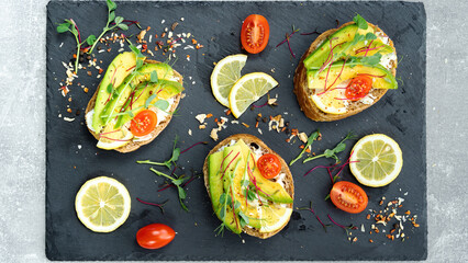 Ciabatta with avocado, cream cheese, cherry tomatoes and microgreens. Snacks with microgreens lie on a black slate serving tray close-up top view. Food photo on a black background.