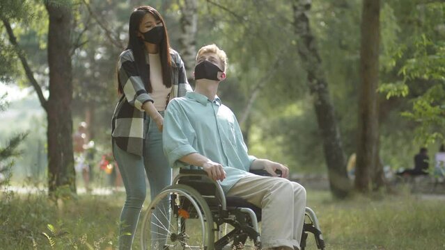 Being in the public park, a handsome asian lady rolls her disabled blonde man in a wheelchair, then they stop, talk a little and decide move away