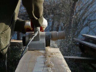 Woodworker planing a long board in a street workshop, a line of sawdust on a texture plank left by an electric planer in the hands of a carpenter working outdoors