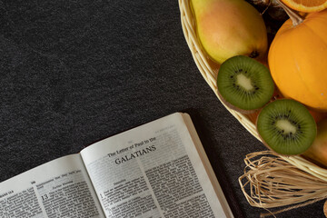 An open Holy Bible book of Galatians with a wicker basket full of various fresh fruits with copy...