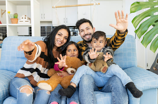 Happy Indian couple and their young children waving hands looking at camera sitting on sofa at home. Joyful family having video call virtual meeting