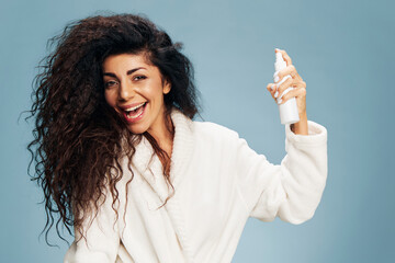 Smiling enjoyed curly Latin lady in bathrobe holding hairspray on hand posing isolated on pastel blue background smiling at camera, feeling excited of effective skincare routine or styling hair