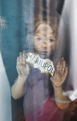 a little Ukrainian girl with sad eyes is standing behind the glass holding a piece of burnt paper with the inscription Mariupol. Children and war.