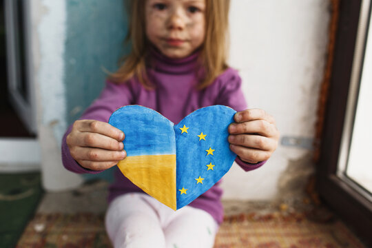 a little Ukrainian girl with a face blackened by fire is sitting on the floor by the window holding a heart painted in the colors of the Ukrainian and European flags. The European Union and Ukraine.