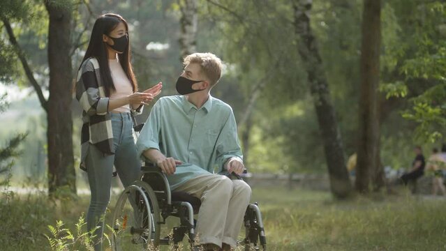 Being in the public park, a handsome asian lady rolls her disabled blonde man in a wheelchair, then they stop, talk a little and decide move away