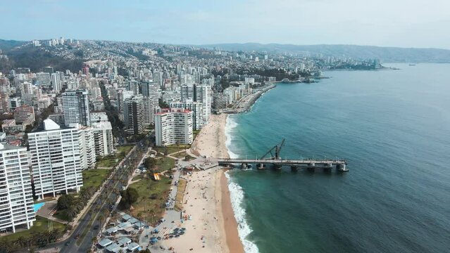 Aerial dolly out of people in El Sol beach shore and turquoise sea near Vergara pier and Vi&ntilde;a del Mar buildings, Valparaiso in background, Chile