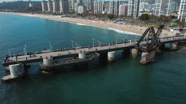 Aerial dolly in of people walking in Vergara pier in turquoise sea near El Sol beach sand shore, Vi&ntilde;a del Mar buildings in background, Chile