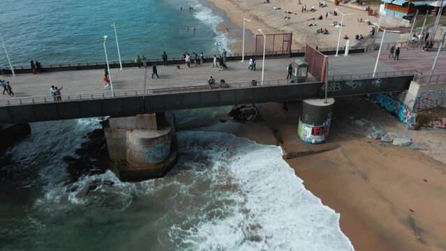 Aerial dolly in of people enjoying Vergara pier and El Sol beach sand shore near turquoise sea, Vi&ntilde;a del Mar buildings in background, Chile