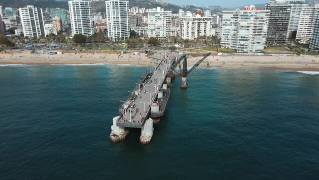 Aerial orbit of touristic Vergara pier and crane in turquoise sea near El Sol beach sand shore, Vi&ntilde;a del Mar buildings in background, Chile