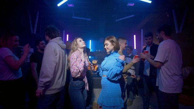 Two girls with a cocktail in their hands are dancing, a crowd of people are dancing around the girls on the dance floor of a nightclub under the light of colored spotlights. Night club