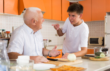 Cheerful teenage boy friendly talking to his elderly grandfather on cozy home kitchen
