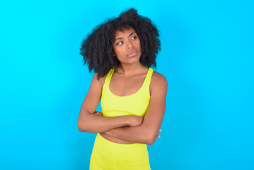 Displeased young woman with afro hairstyle in sportswear against blue background with bad attitude, arms crossed looking sideways. Negative human emotion facial expression feelings.