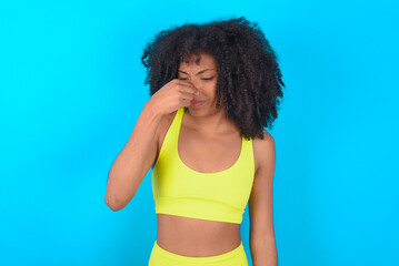 young woman with afro hairstyle in sportswear against blue background, holding his nose because of...