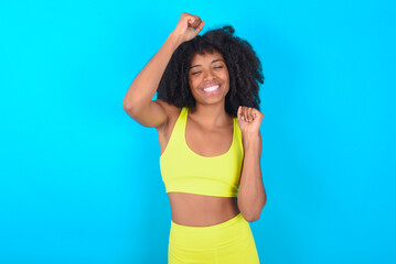 Fototapeta na wymiar Attractive young woman with afro hairstyle in sportswear against blue background celebrating a victory punching the air with his fists and a beaming toothy smile.