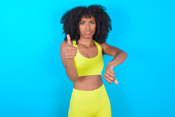 Fototapeta na wymiar young woman with afro hairstyle in sportswear against blue background feeling unsure making good bad sign. Displeased and unimpressed.