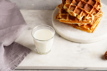 Sweet breakfast - fresh homemade belgian waffles with honey and powdered sugar stacked on white round marble board and small glass with milk