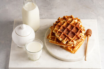 Sweet breakfast - fresh homemade belgian waffles with honey and powdered sugar stacked on white round marble board and small glass with milk