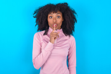 Surprised young woman with afro hairstyle in technical sports shirt against blue background makes silence gesture, keeps finger over lips and looks mysterious at camera