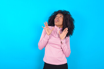 young woman with afro hairstyle in technical sports shirt against blue background keeps palms forward and looks with fright above on ceiling tries to defense herself from invisible danger opens mouth.
