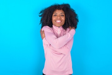 Fototapeta premium Charming pleased young woman with afro hairstyle in technical sports shirt against blue background embraces own body, pleasantly feels comfortable poses. Tenderness and self esteem concept