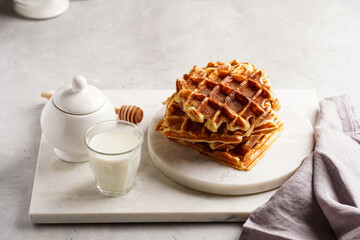 Sweet breakfast - fresh homemade belgian waffles with honey and powdered sugar stacked on white round marble board and small glass with milk