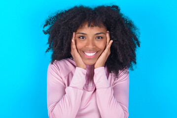 Happy young woman with afro hairstyle in technical sports shirt against blue background touches both cheeks gently, has tender smile, shows white teeth, gazes positively straightly at camera,