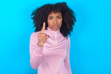 young woman with afro hairstyle in technical sports shirt against blue background frustrated and pointing to the front