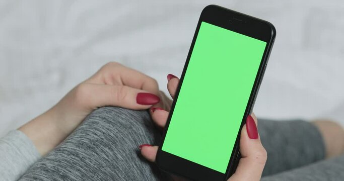 Woman sitting on the bed and holding smartphone with pre-keyed green screen. Mock up and chroma key. Technologies and people concept. Close up