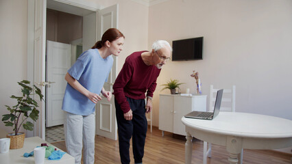 A woman physiotherapist turns on a video with exercises on a laptop and explains to a pensioner how to perform them