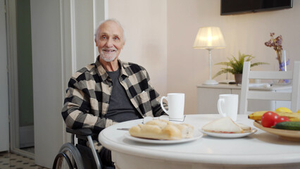 An old man in a wheelchair sits at a dining table and looks at the camera