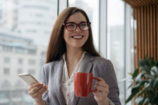 Beautiful smiling businesswoman wearing stylish eyeglasses using mobile phone holding cup of coffee looking away in office. Coffee break concept 