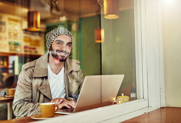 Hanging out at his favorite cafe. Portrait of a stylish young man smiling while drinking a coffee and using a laptop in a cafe.