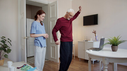 A physiotherapist watches as an old man swings his arms. He repeats the exercises that he sees on...