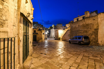 A quiet street in the ancient city of Matera, Italy, at night, with a car parked and a dog standing under a lamp in the distance. - Powered by Adobe