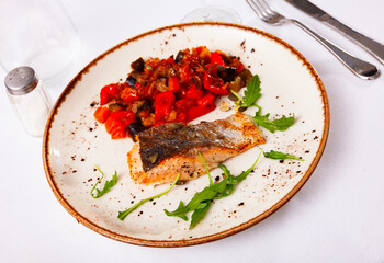 Steamed salmon fillet with eggplants and tometoes served on plate and decorated with arugula, over white background.