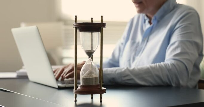 Mature businesswoman sit at desk turns hourglass over, while working on laptop, close up focus on sand glass. Measuring time., deadline, urgent task, effective productive workflow, countdown concept